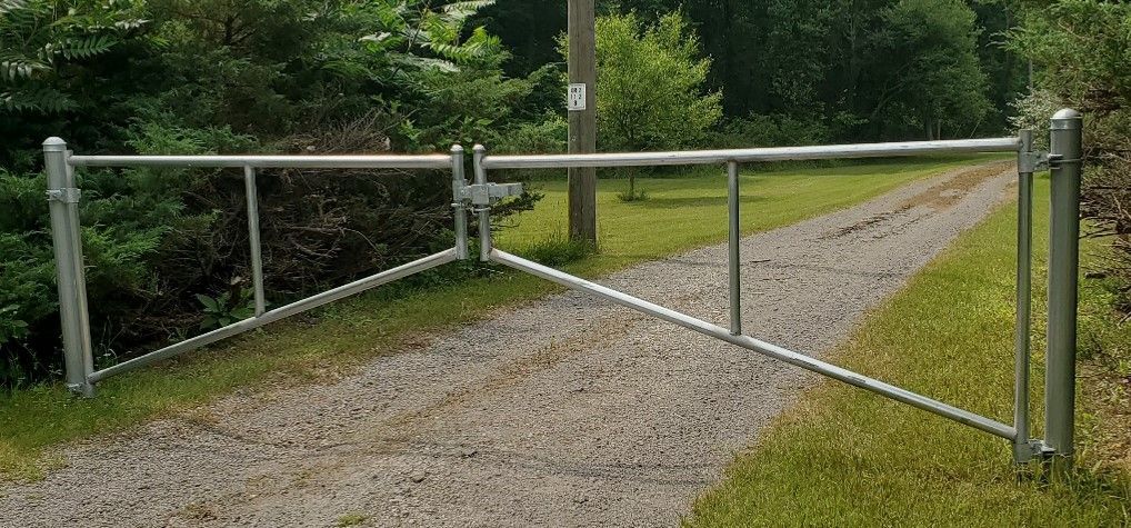 A metal gate, partly open, blocks a gravel driveway leading into a wooded area.