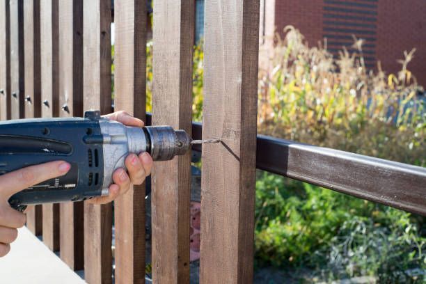 Man drilling hole in wooden fence with electric drill outdoors on sunny day for home improvement.