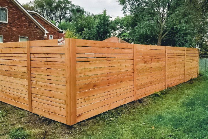 Wooden horizontal slat fence in a backyard. Brown wood, green grass, brick house in background.