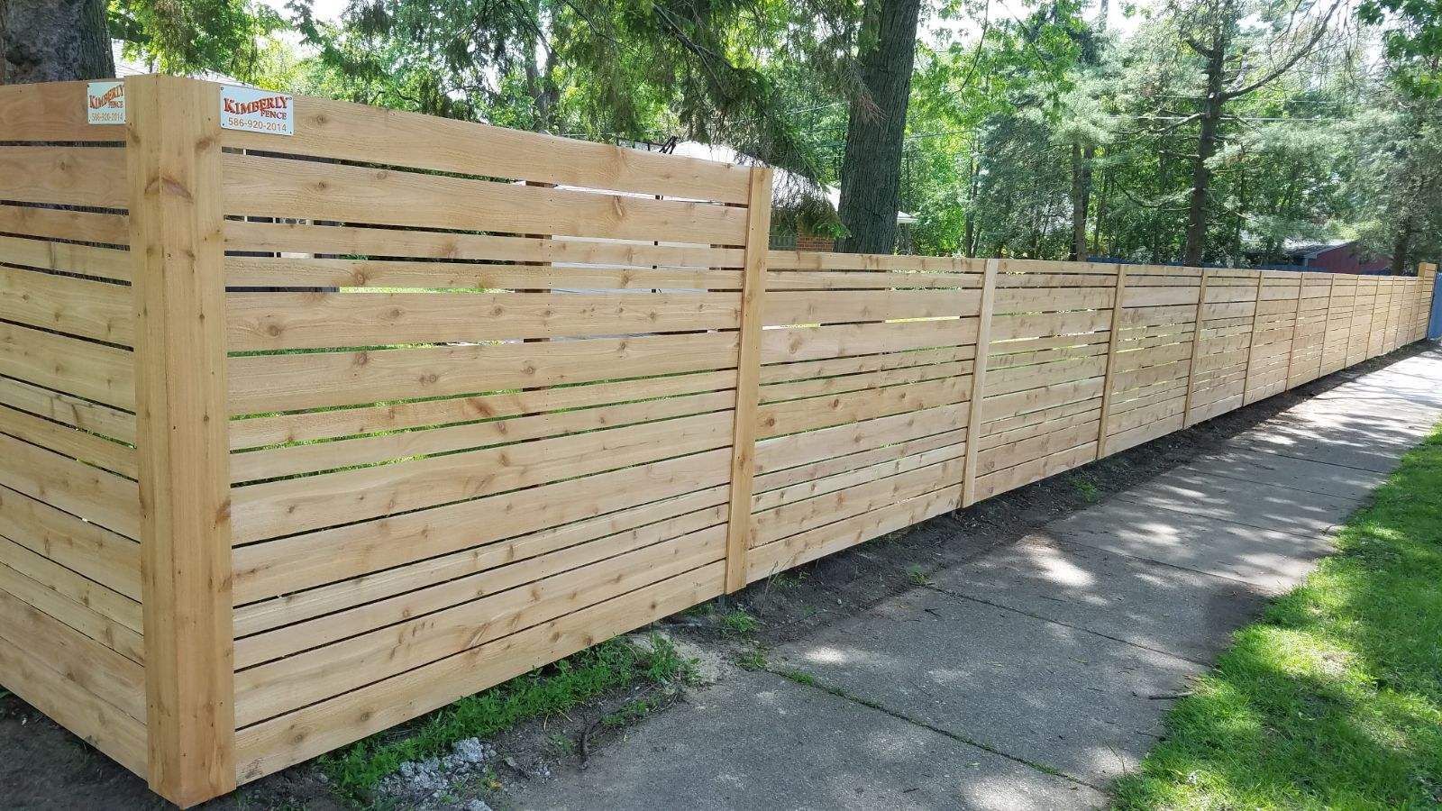 Wooden horizontal slat fence along a sidewalk, with green grass and trees visible.
