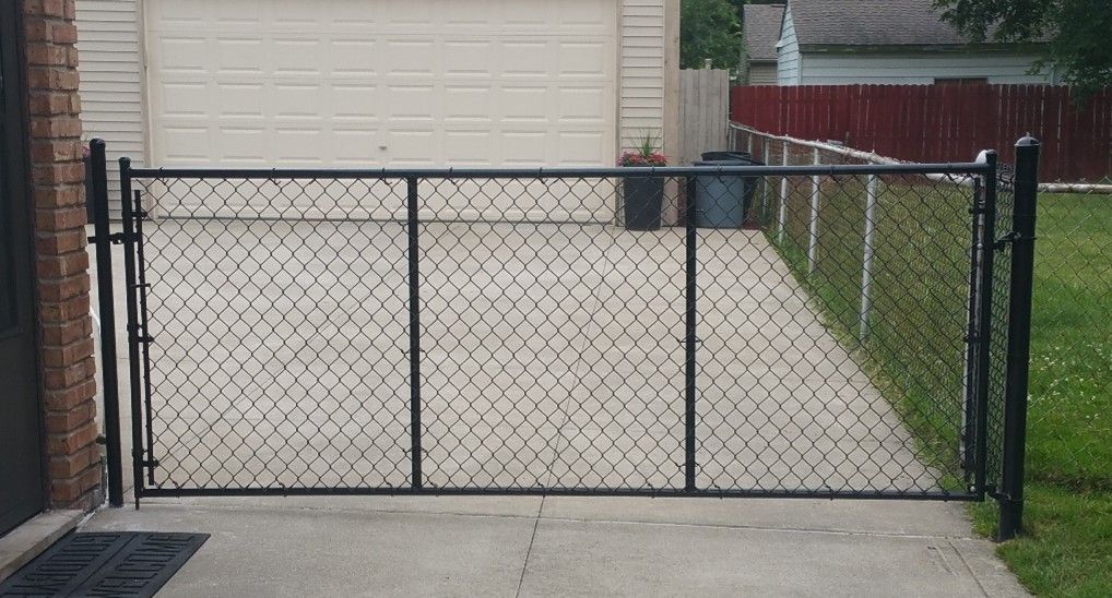Black chain-link gate on a concrete driveway leading to a garage, with a red fence visible in the background.