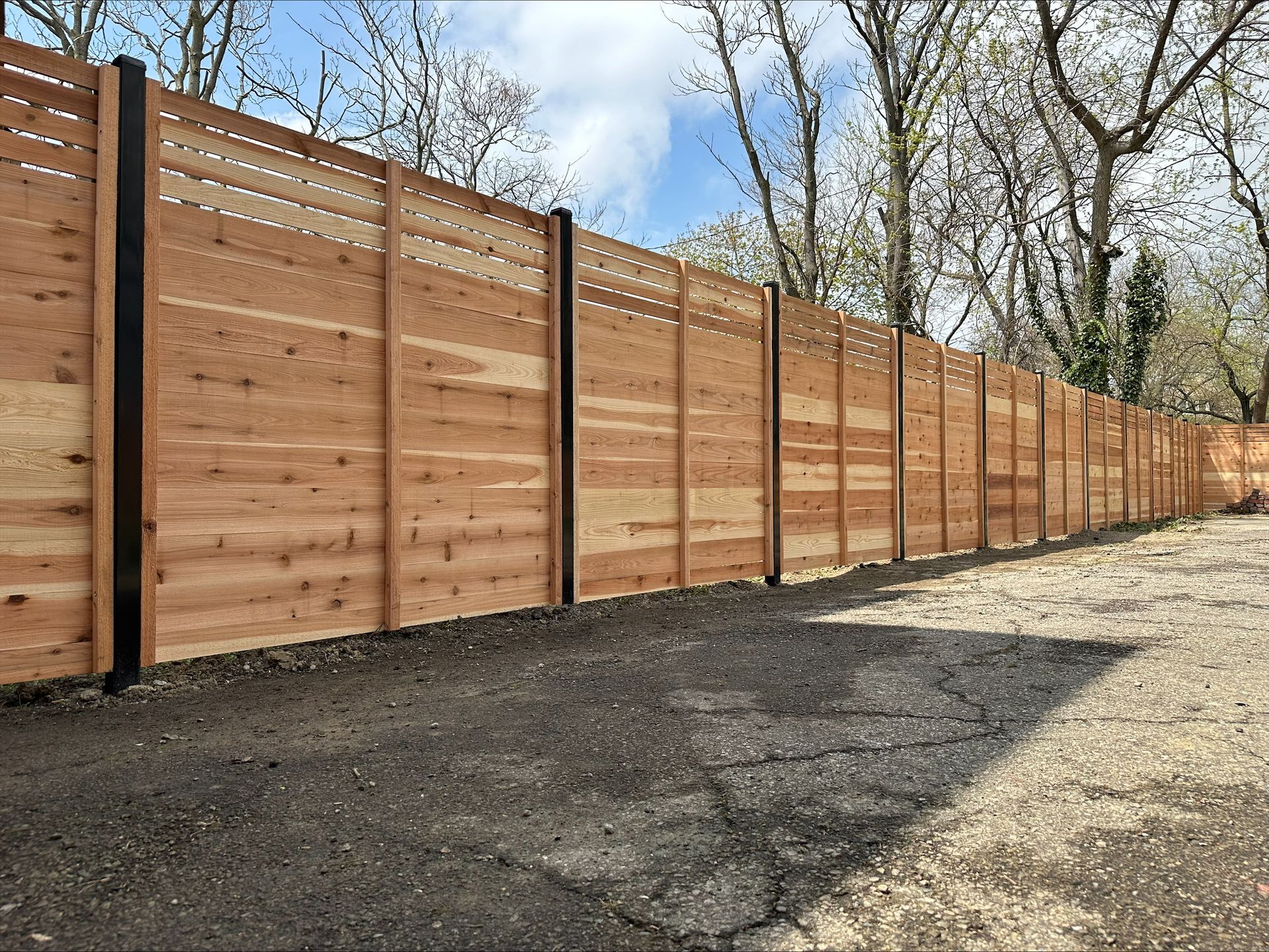 Wooden fence, brown horizontal planks with black posts, in a gravel setting, trees in background.