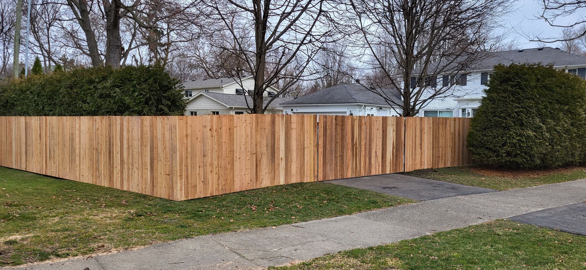 Wooden fence bordering a yard with grass, sidewalk, and trees; houses in the background.