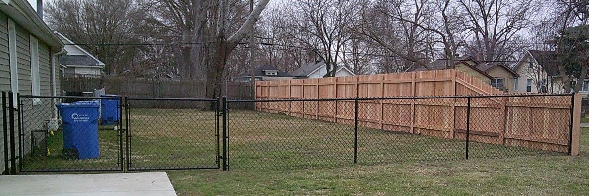 A backyard with a wooden fence and a chain-link fence, with two blue trash cans near a house.