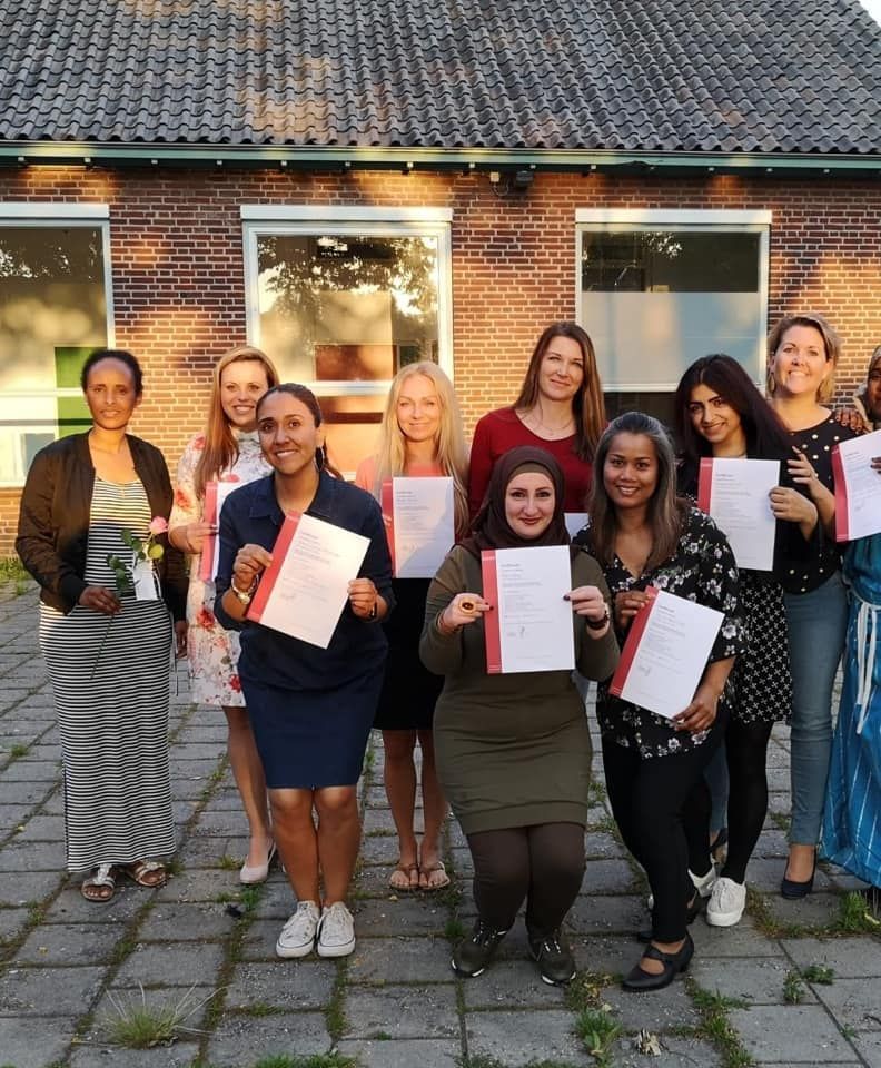Een groep vrouwen staat buiten met certificaten in de hand en lacht bij een bakstenen gebouw.