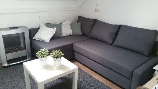 Grijze hoekbank met kussens, witte salontafel en zilverkleurige elektrische kachel in een kleine woonkamer.