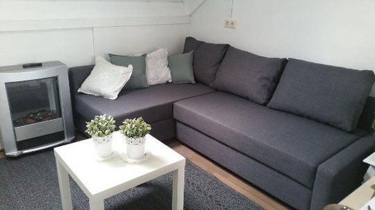 Grijze hoekbank met kussens, witte salontafel en zilverkleurige elektrische kachel in een kleine woonkamer.