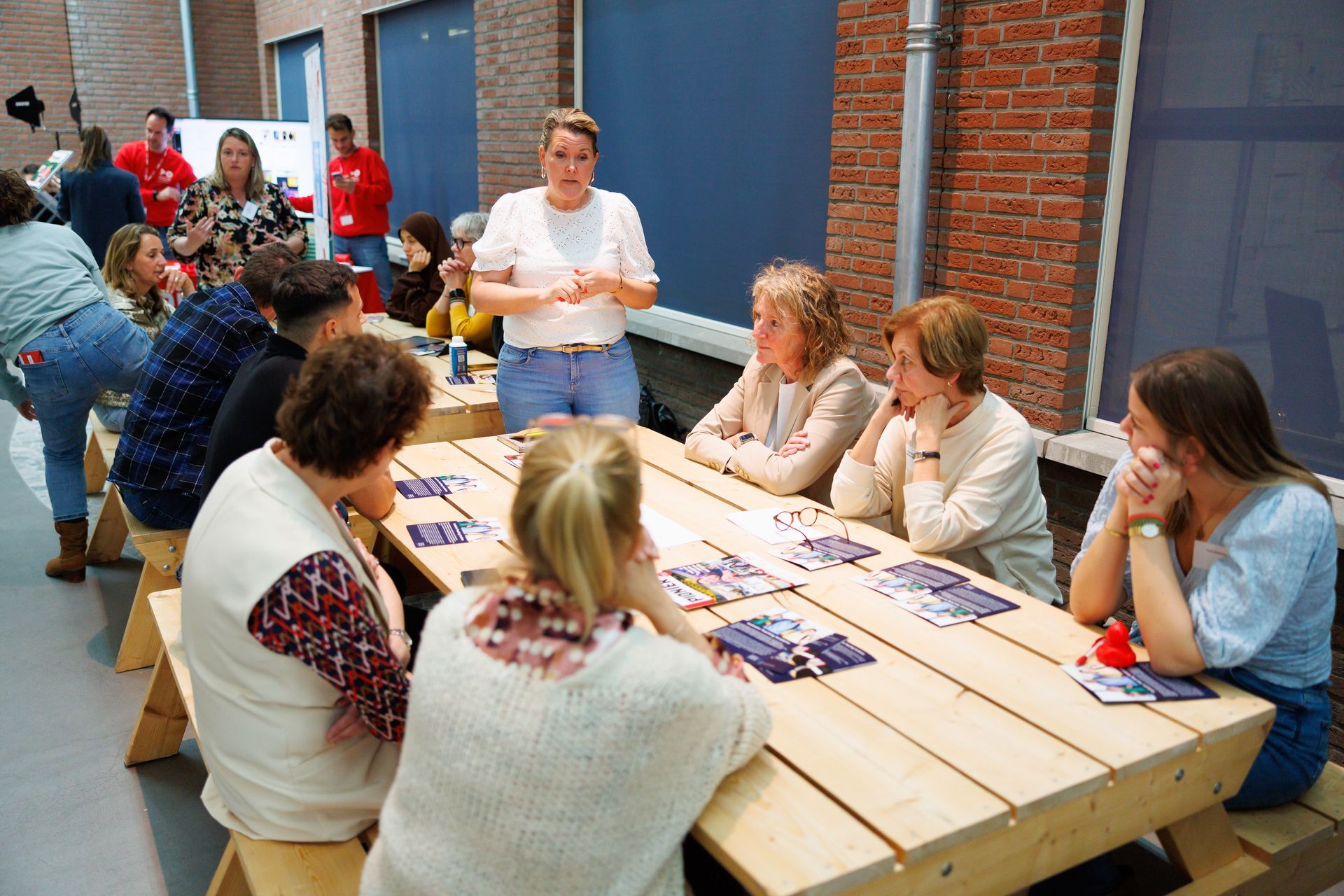 Mensen zitten aan een tafel en luisteren naar Femke die gebaart, terwijl anderen toekijken. Binnen, met een bakstenen muur op de achtergrond.