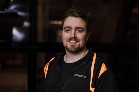Smilende mann i svart jakke med oransje detaljer mot en mørk innendørsbakgrunn