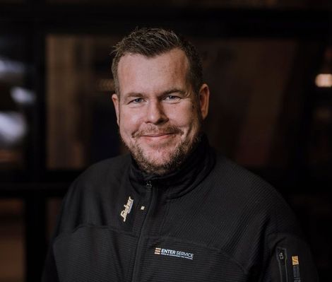 Smilende mann i en svart jakke mot en mørk, uskarp bakgrunn