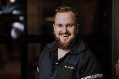 Smilende person i en svart jakke som står innendørs mot en mørk, uskarp bakgrunn