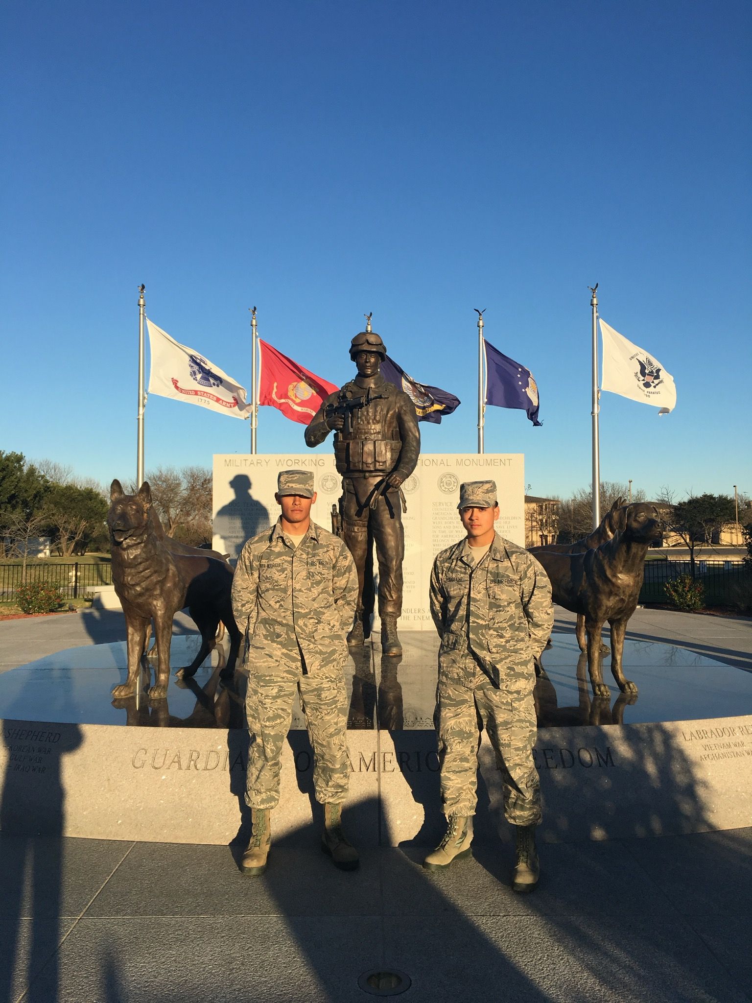 Two soldiers in camo stand before a military memorial with statues and flags on a sunny day.
