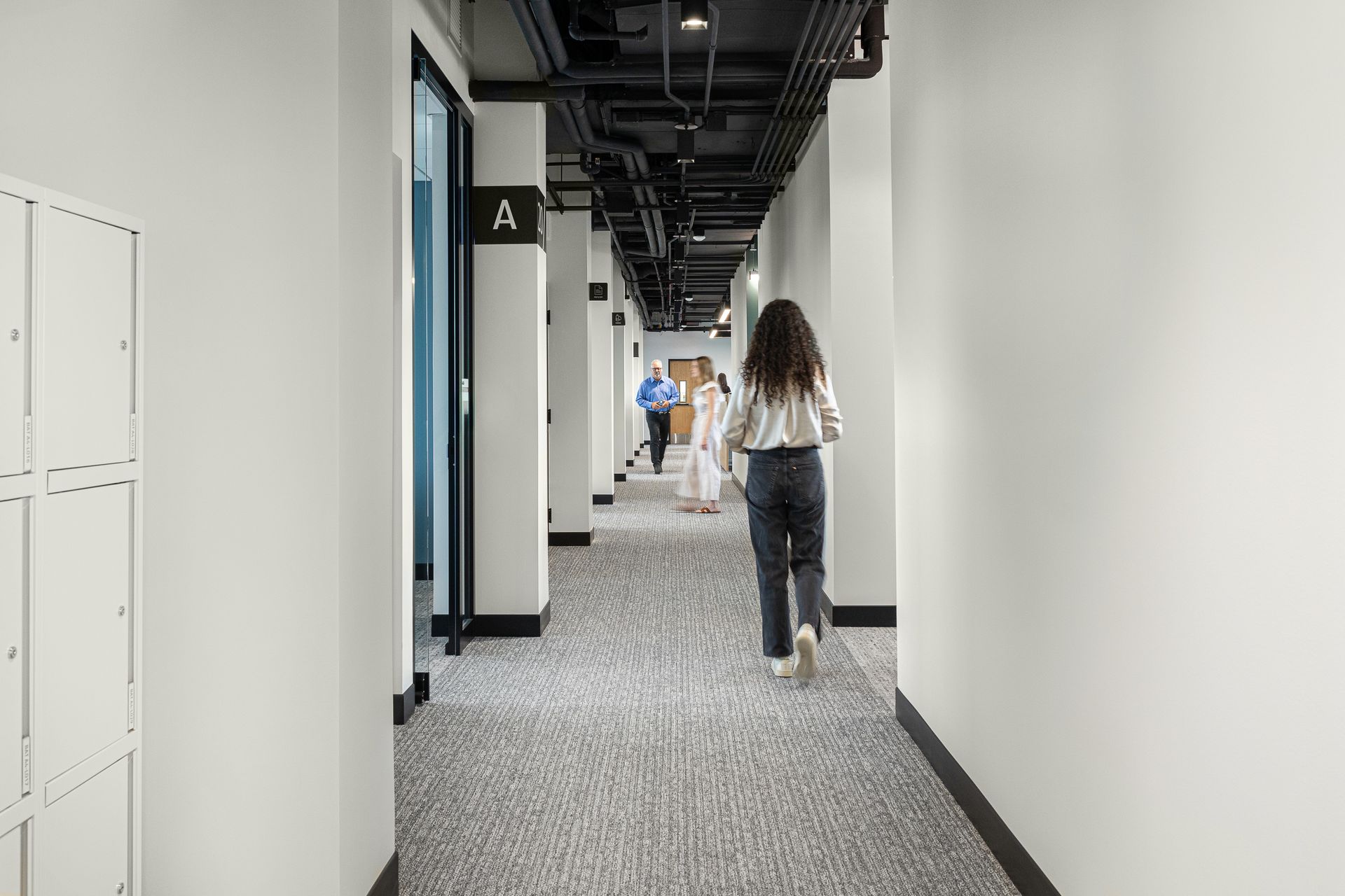 Woman walking down a long, white hallway with offices on the left and a few people further down.