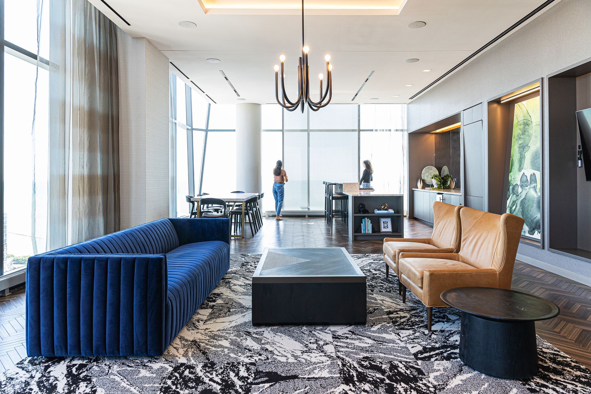 Modern living room with blue sofa, leather chairs, and black chandelier; two people at a counter.