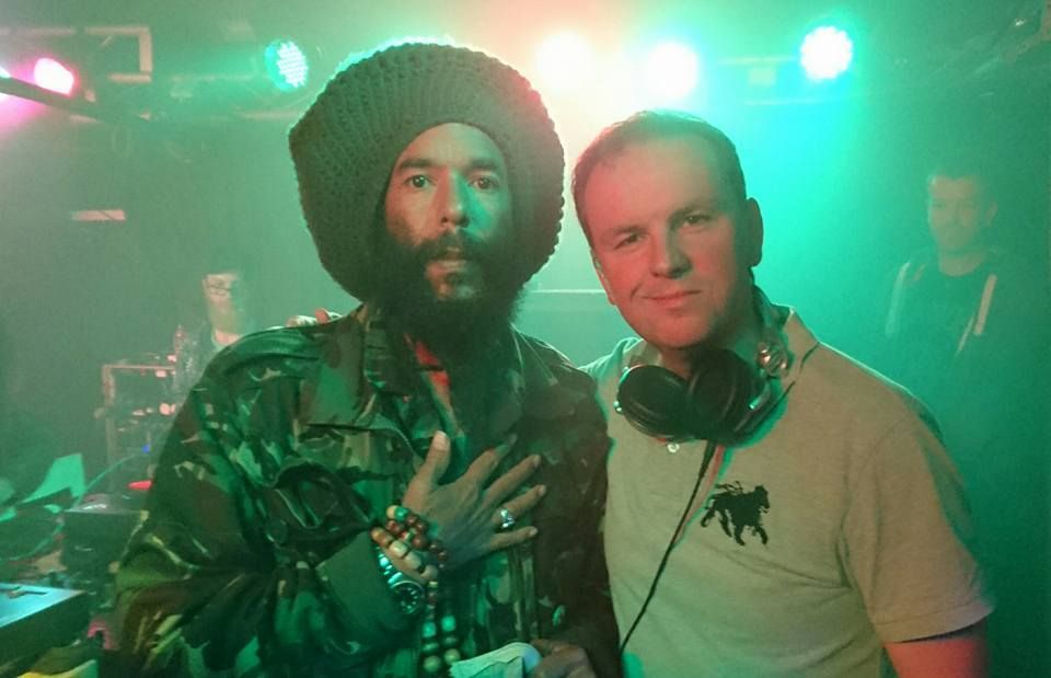 Two people posing in a neon-lit club, one with headphones around his neck and bright stage lights behind them