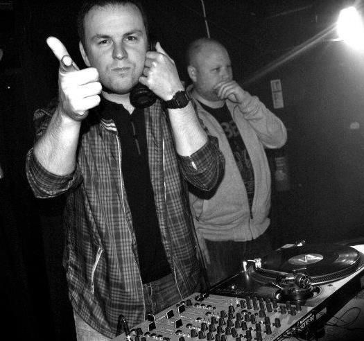 DJ at a club booth giving a thumbs-up, with another man behind him and turntables in the foreground