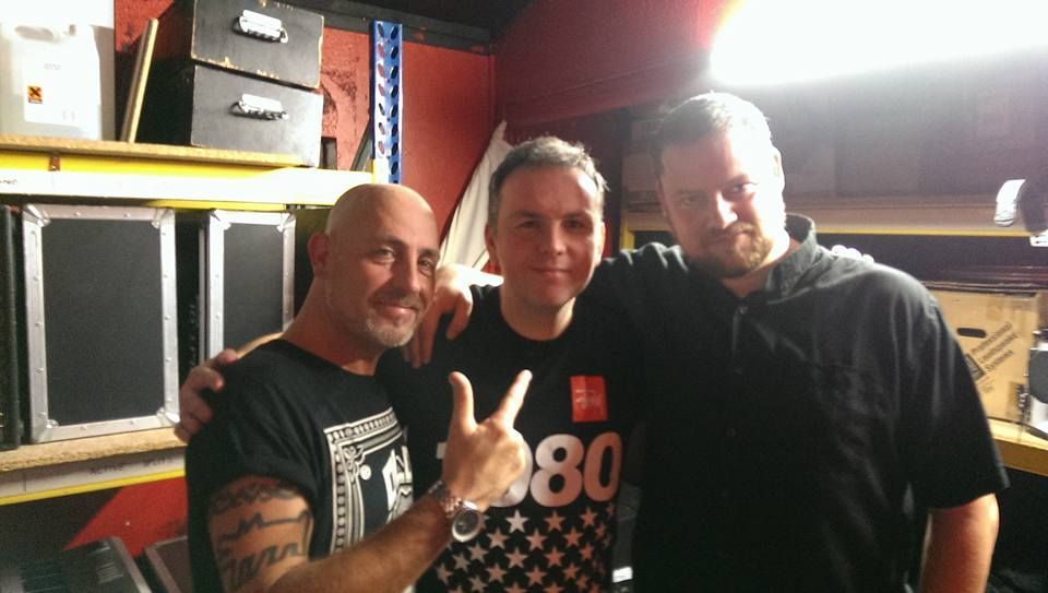 Three people posing together indoors, one flashing a peace sign, in a cluttered room.