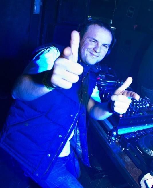 Smiling DJ in blue lighting giving two thumbs up beside a mixing console