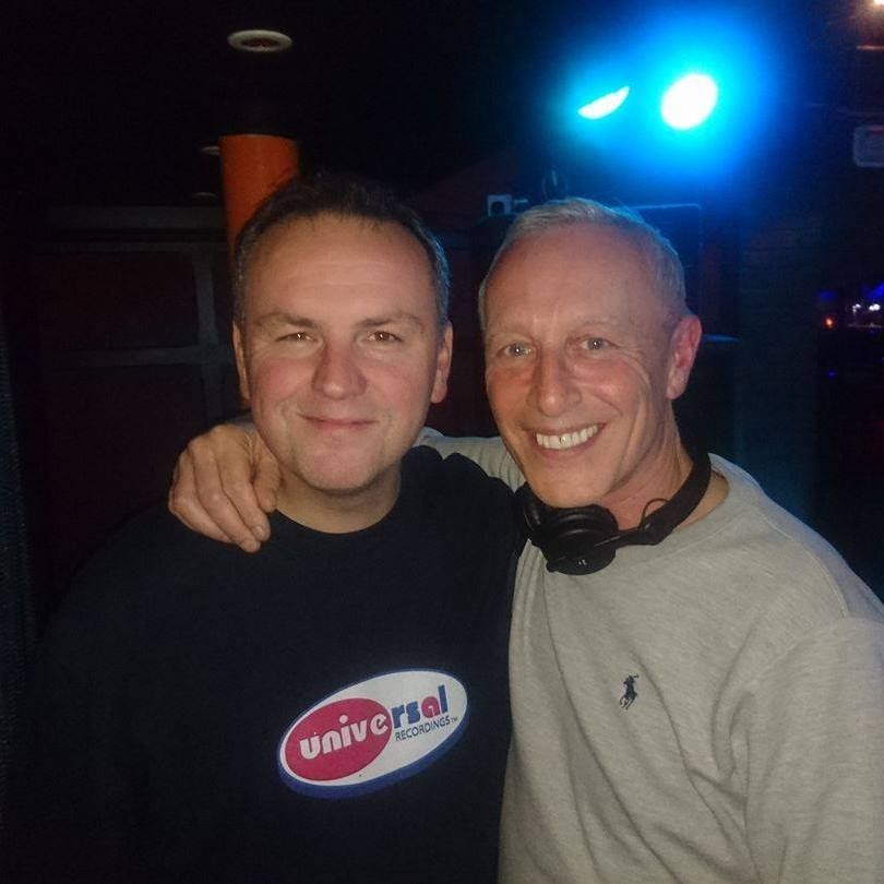 Two smiling people posing together in a dim club with blue lights and headphones around one neck