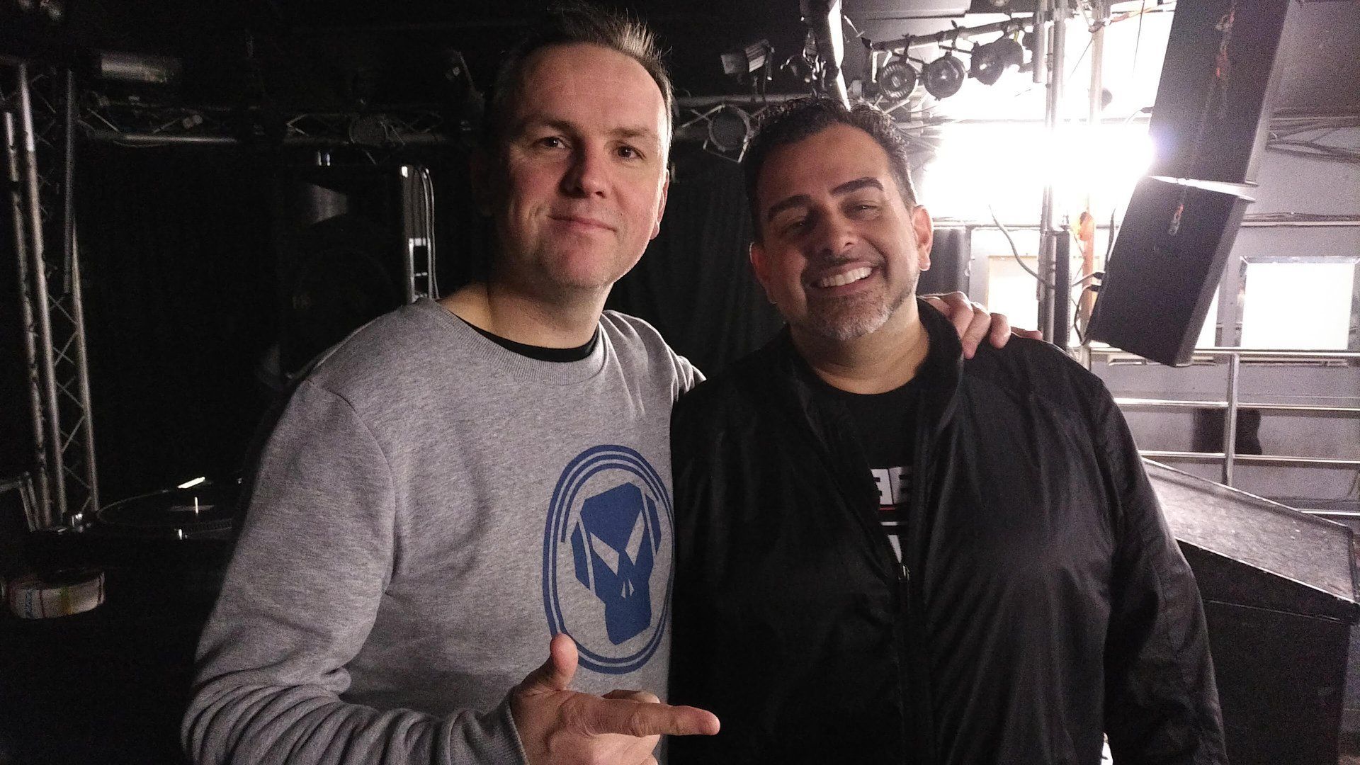 Two smiling men standing together in a dimly lit venue, with stage lights glowing behind them.