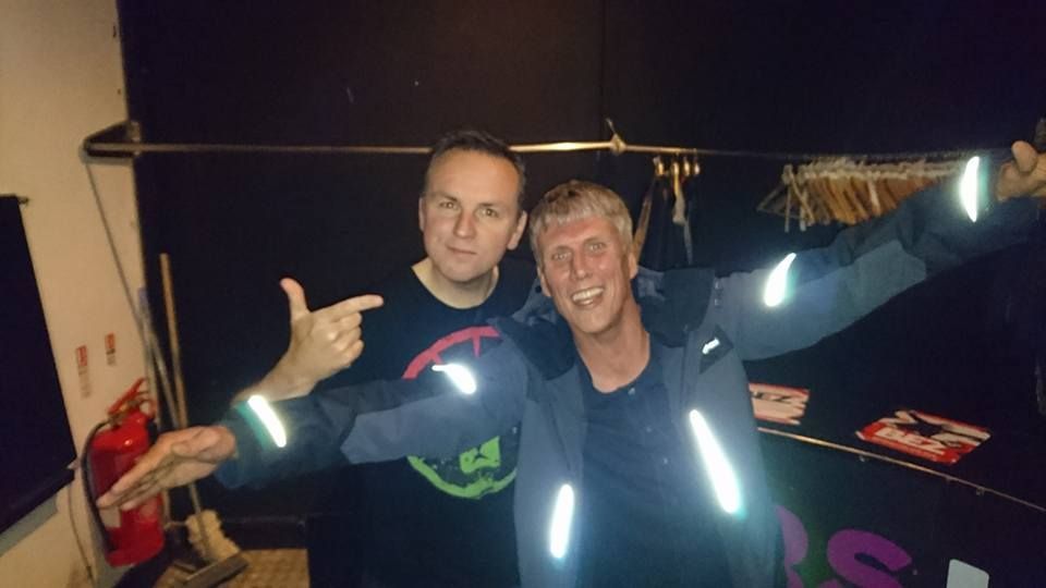 Two people smiling in a dim indoor space, one pointing at the other with bright reflective stripes on their clothing
