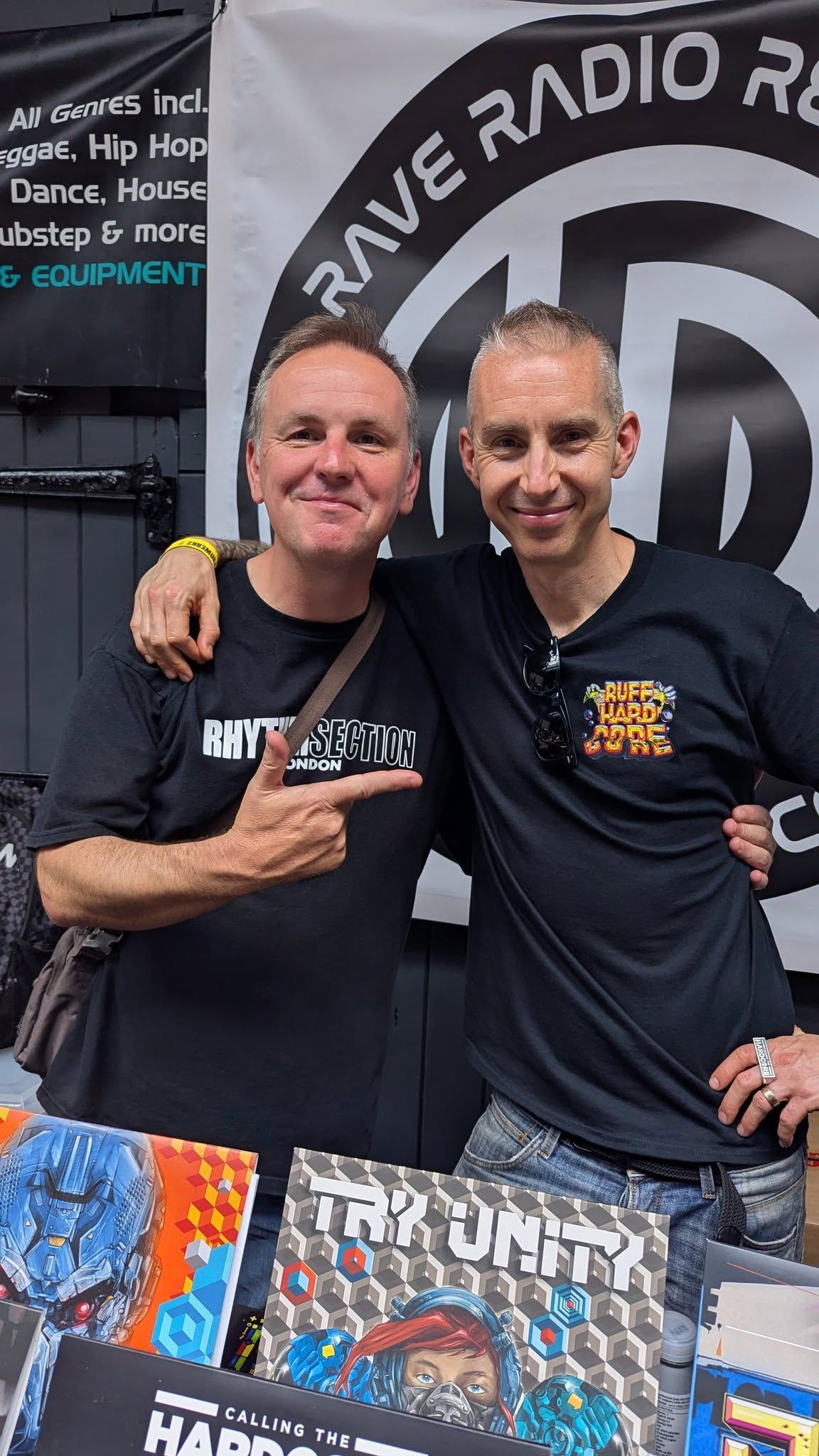 Two men posing together at a radio station booth with event merchandise displayed in front of them.