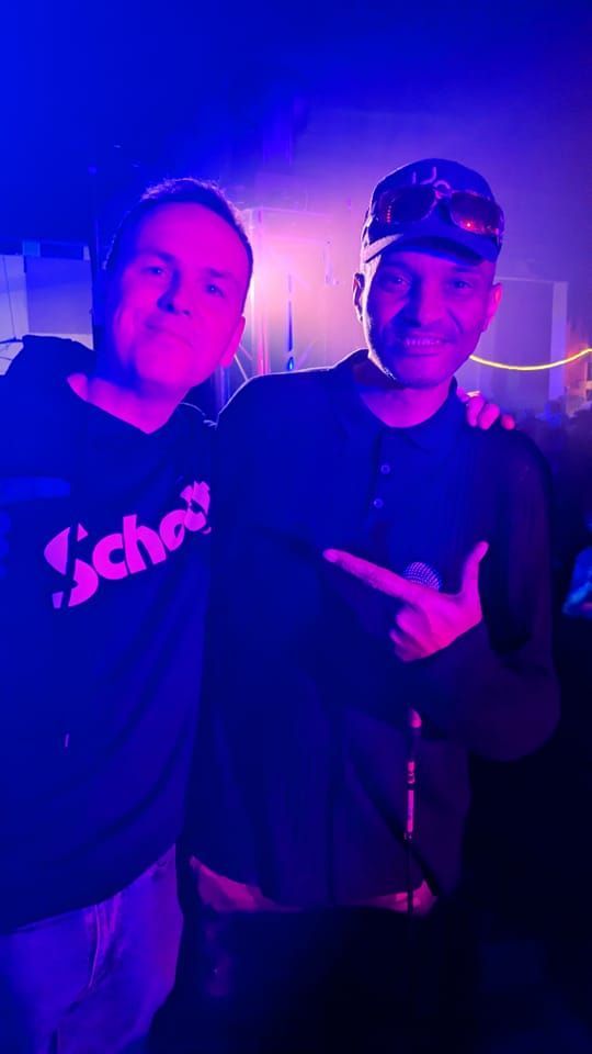 Two people posing together under purple-blue party lighting, one pointing at the other.