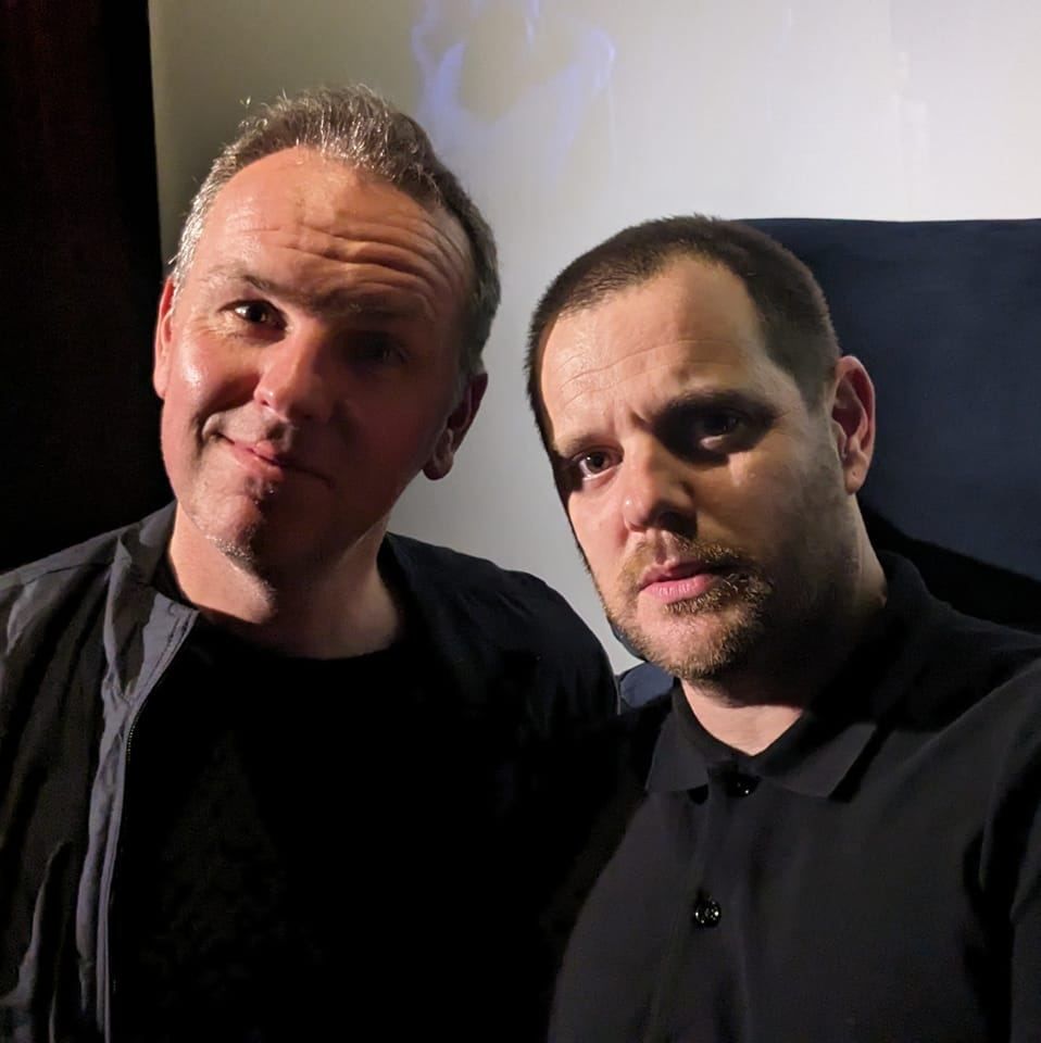 Two men posing for a selfie in dim light, wearing dark shirts and looking at the camera.