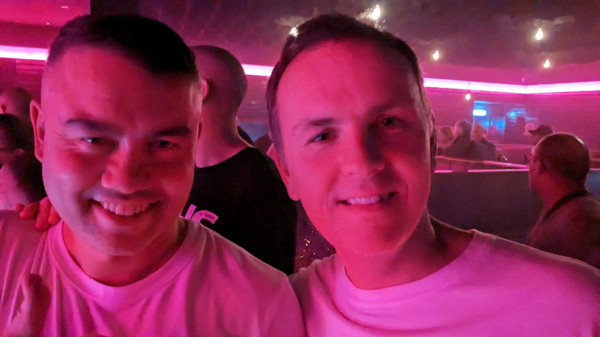 Two people smiling in a pink-lit nightclub, with a crowded background.