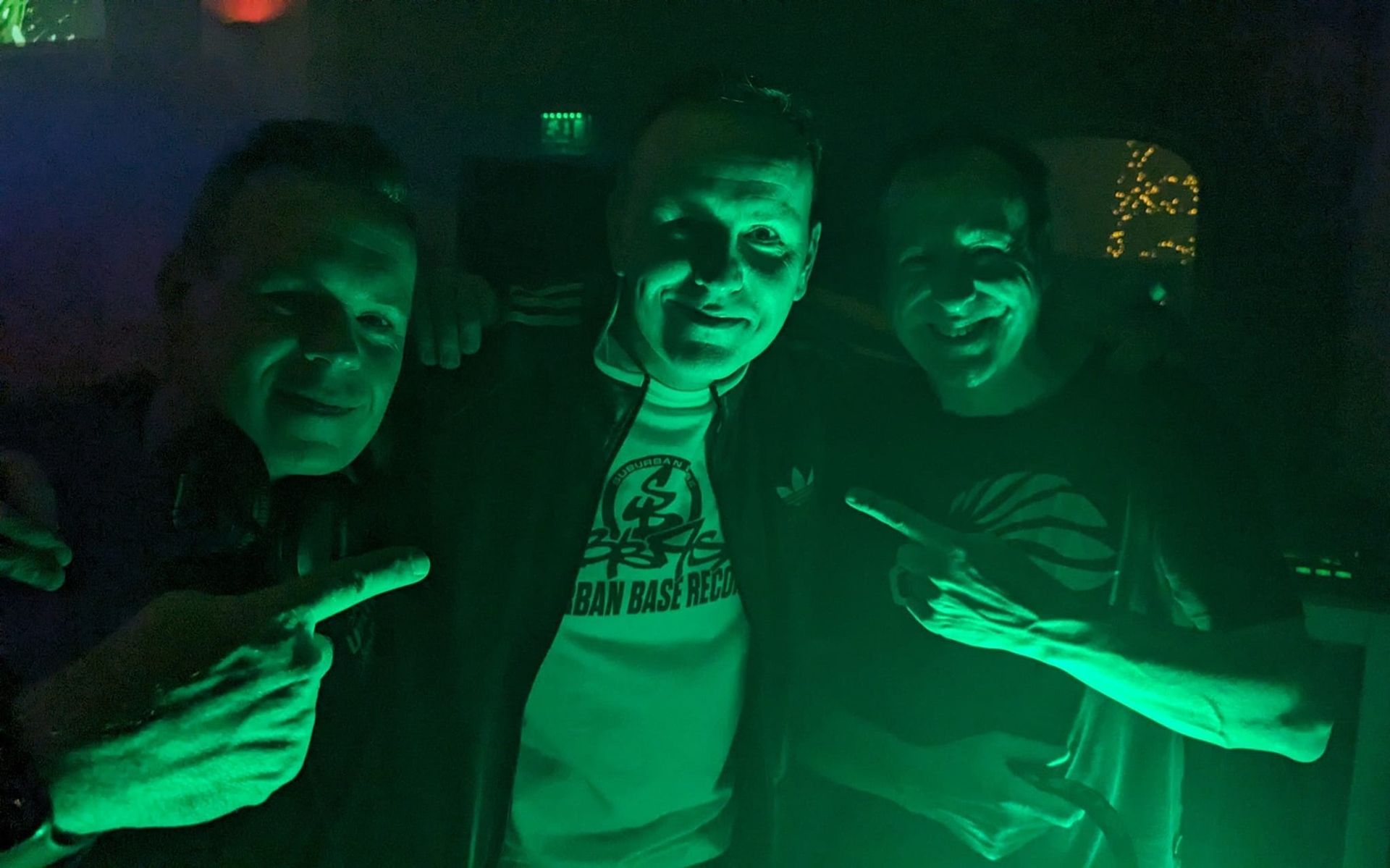 Three people posing in green club lighting, smiling and flashing hand signs.