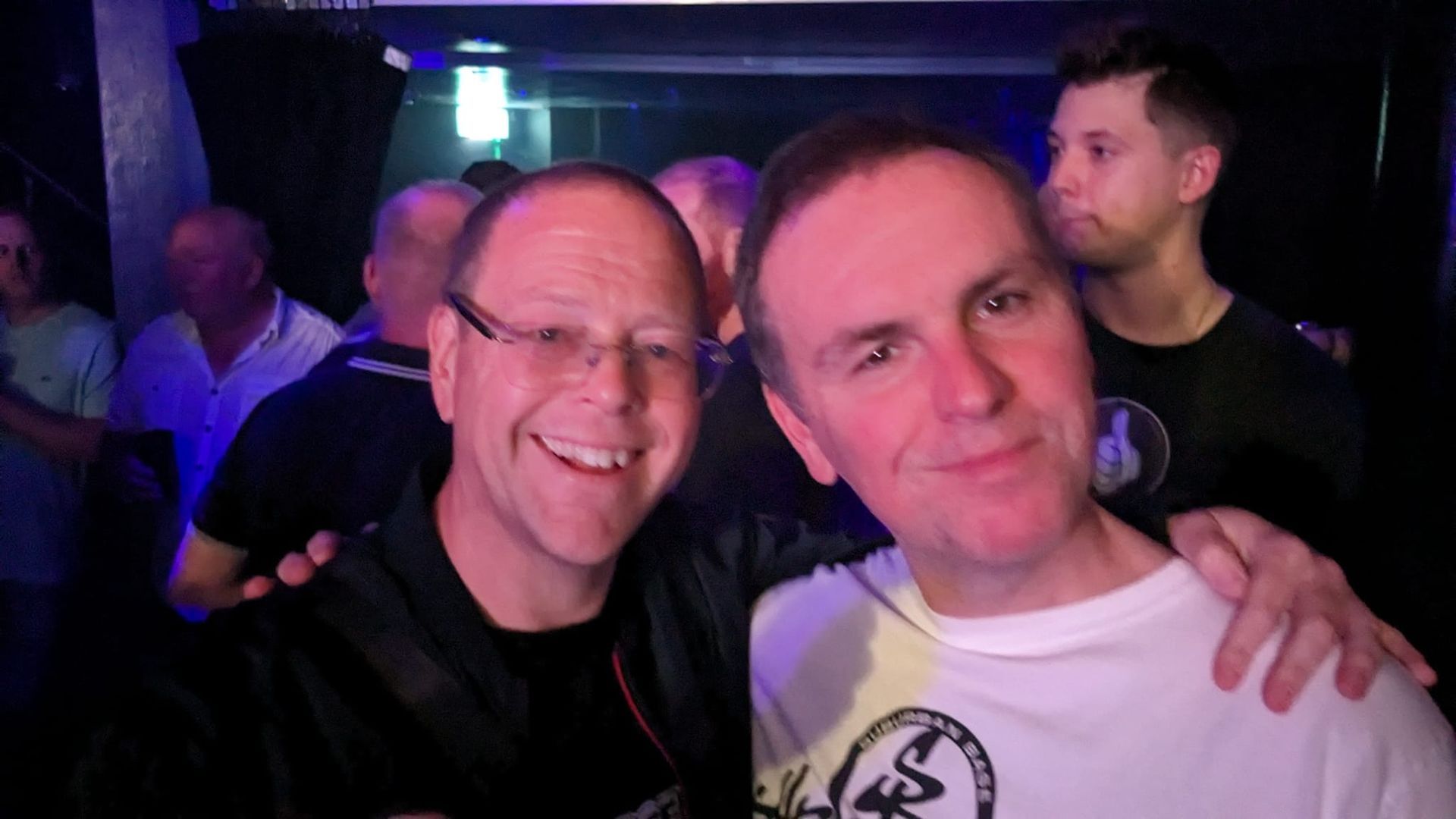 Two smiling men posing together in a dimly lit nightclub, one with an arm around the other.