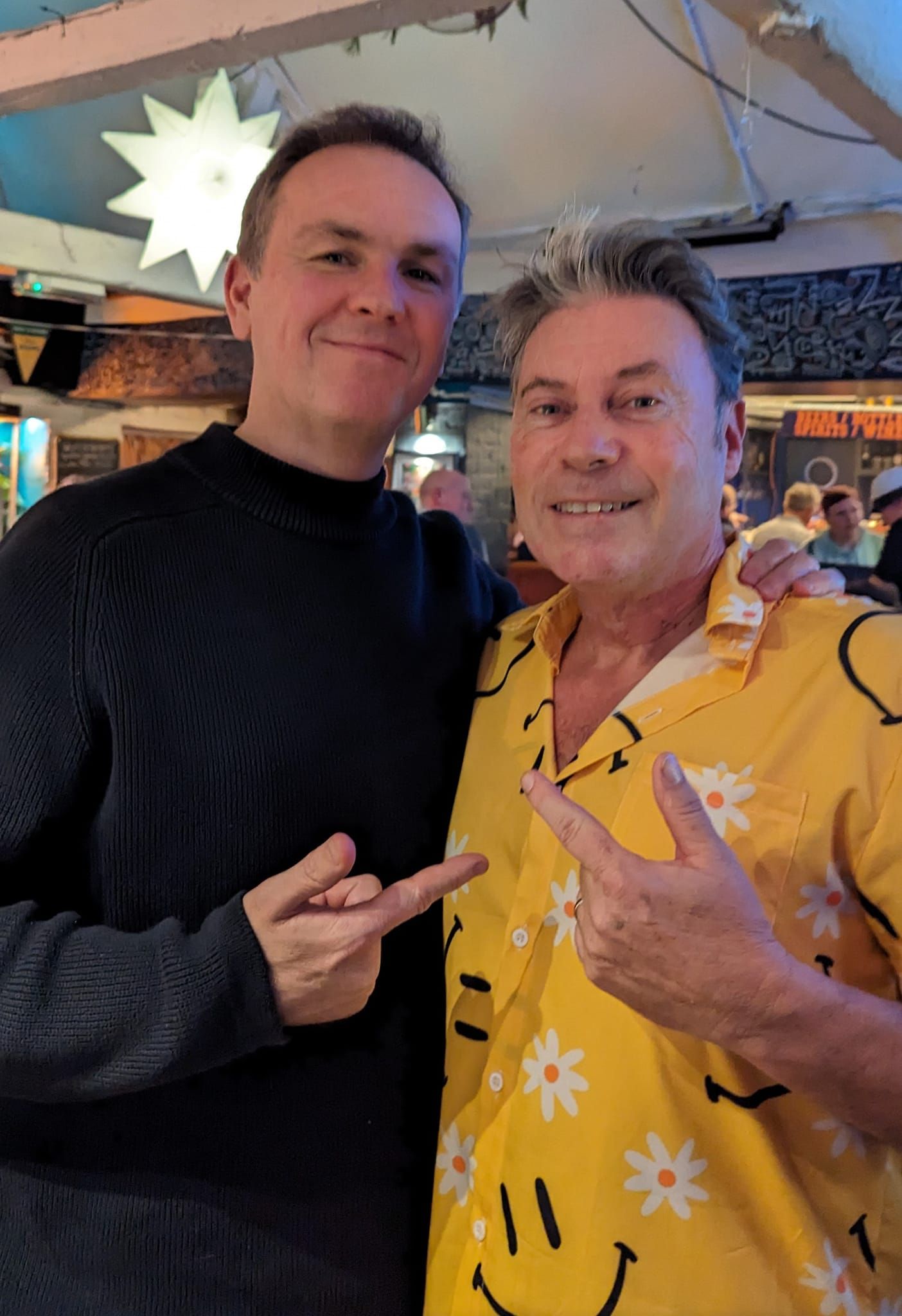 Two smiling men posing indoors, one in a black sweater and one in a yellow shirt, both making peace signs.