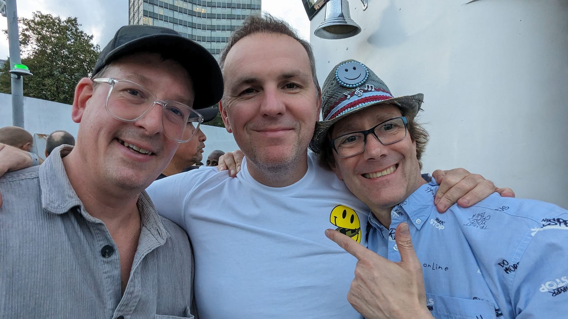 Three smiling people posing close together outdoors, with one wearing a cap and another wearing glasses and a patterned hat.