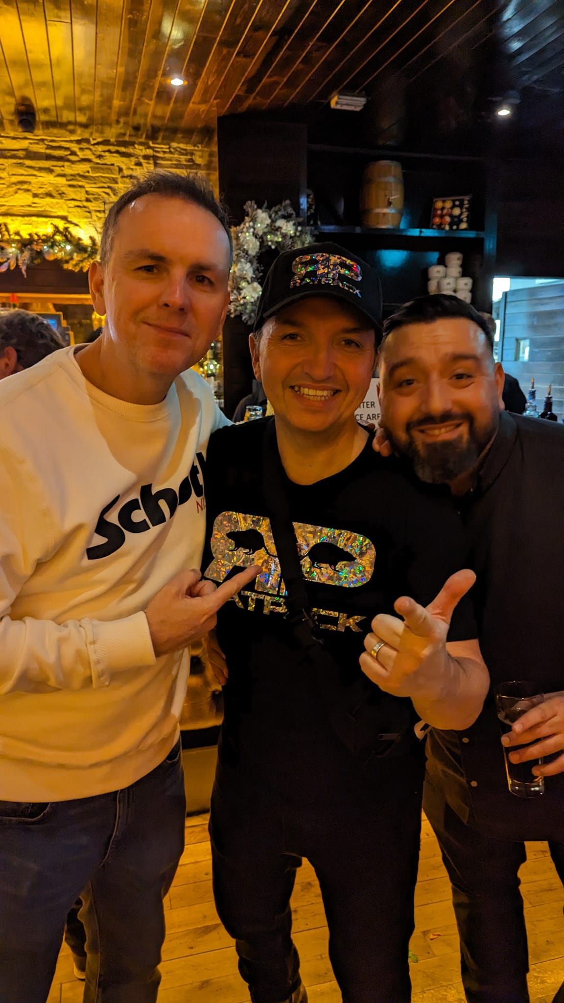 Three people smiling in a dim bar, posing with arms around each other under warm ceiling lights.