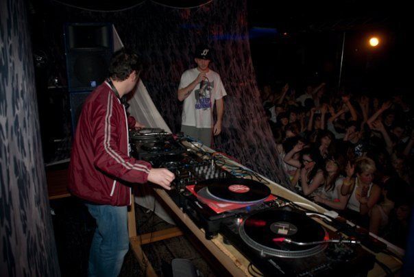 DJ performing at a club booth with vinyl turntables, facing a cheering crowd under dim lights
