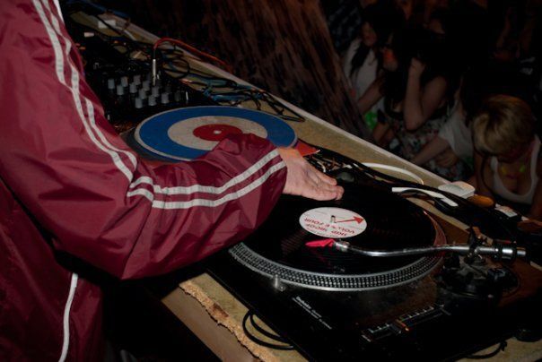 DJ in a red jacket using a turntable with a blue-and-red record in a dim club setting