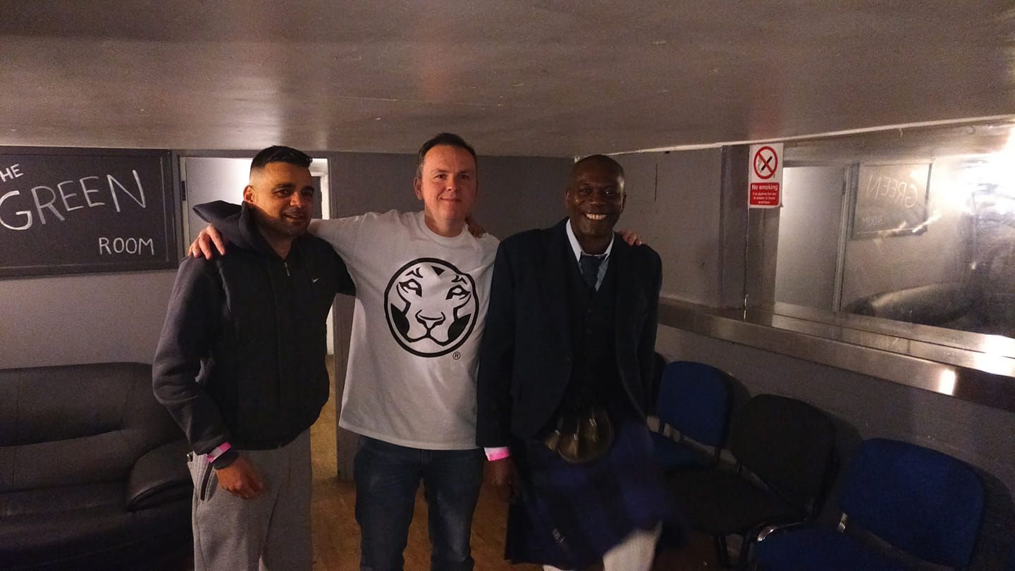 Three men posing together in a dim hallway, with a green room sign and blue chairs nearby.