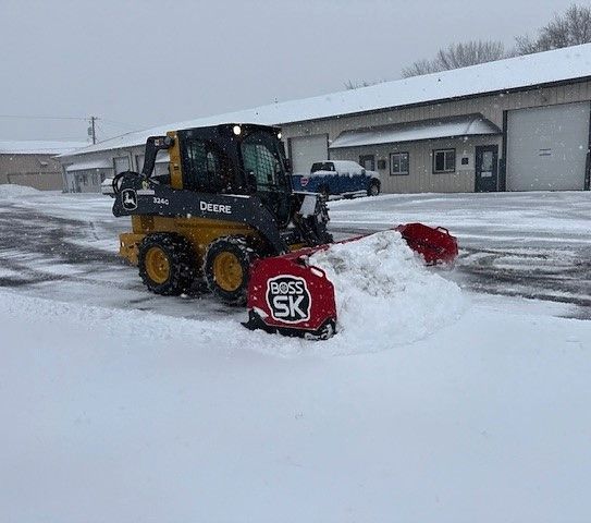 Skid steer with snow plow clearing snow in front of a building during a snowfall.