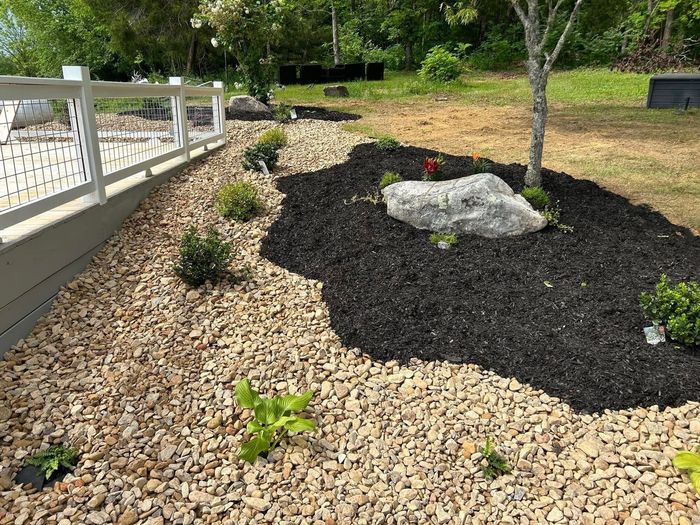 Landscape bed with black mulch, tan gravel, a small tree, and a white railing by the path
