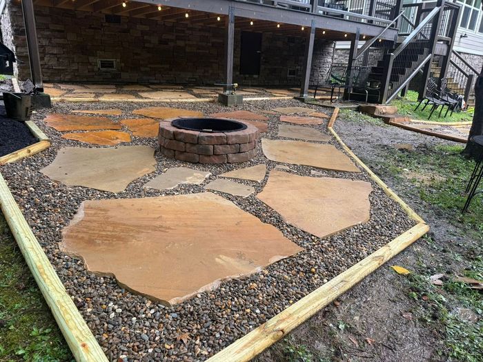 Stone patio with a circular fire pit, bordered by gravel and a wooden frame near a house.