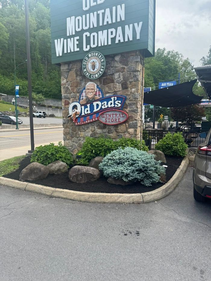 Roadside sign for Mountain Wine Company and Red Dogs, with rocks, shrubs, and parked cars nearby