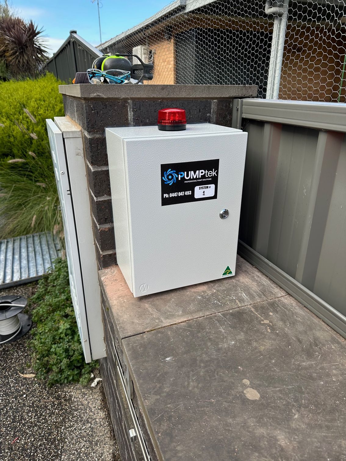 White utility cabinet with a red beacon on a narrow outdoor concrete walkway beside a fence and brick wall — PUMPtek in Grovedale, VIC