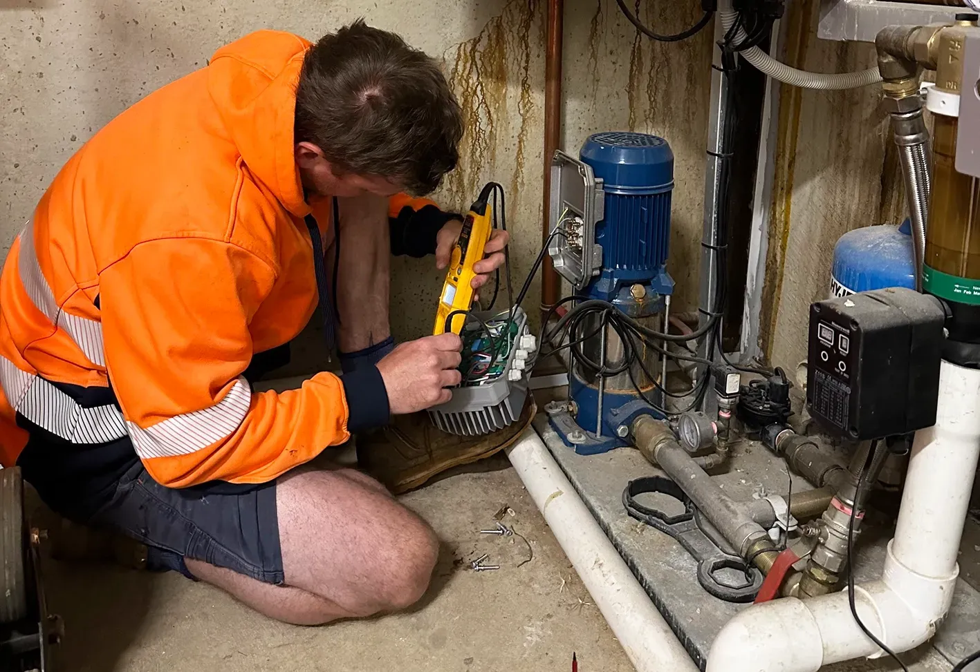 Technician in orange jacket kneels by basement pumps, inspecting plumbing with a flashlight and tool  — PUMPtek in Bendigo, VIC