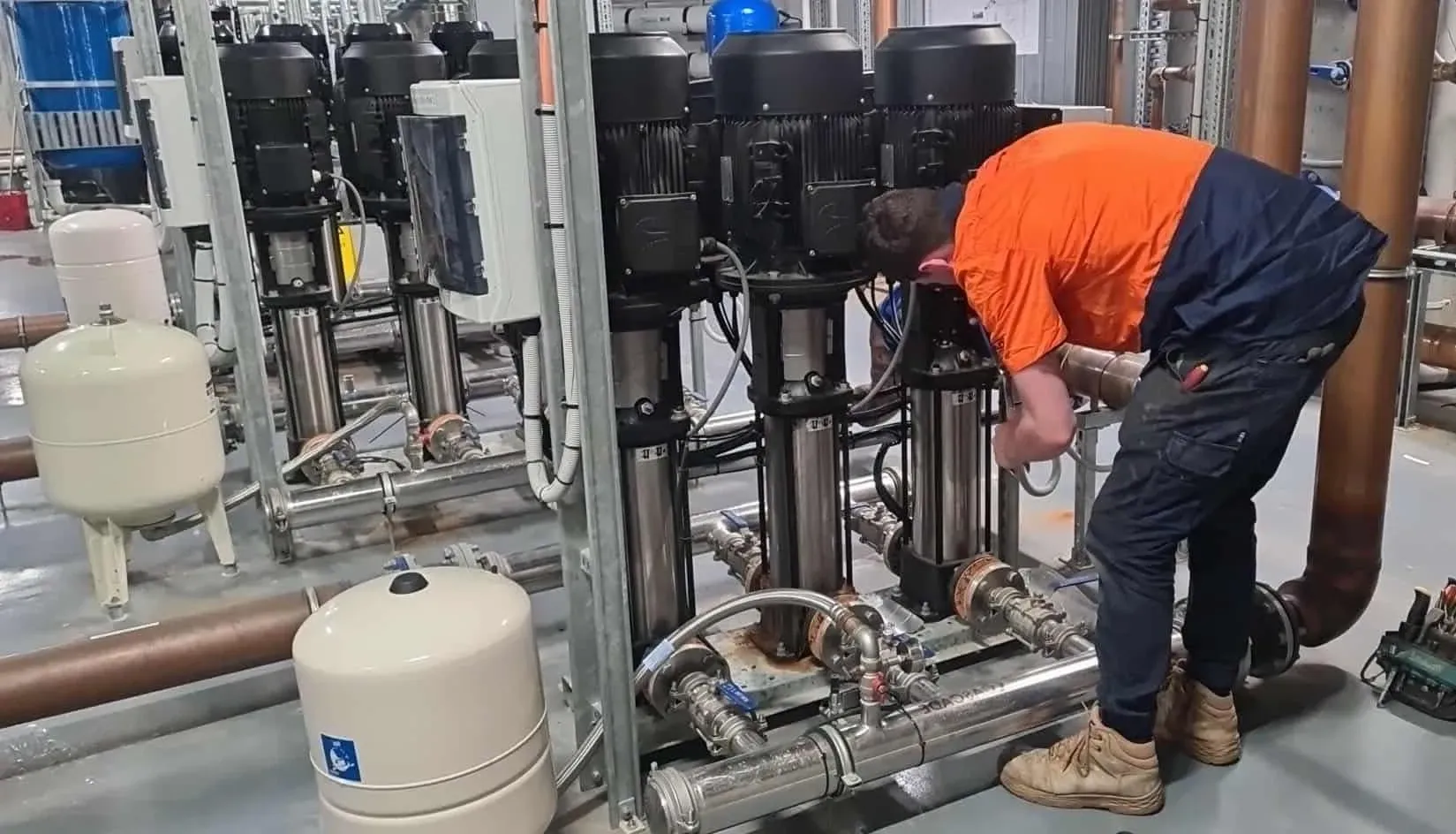 Worker in orange and navy uniform bends over pipes and pumps in an industrial plant — PUMPtek in Bendigo, VIC