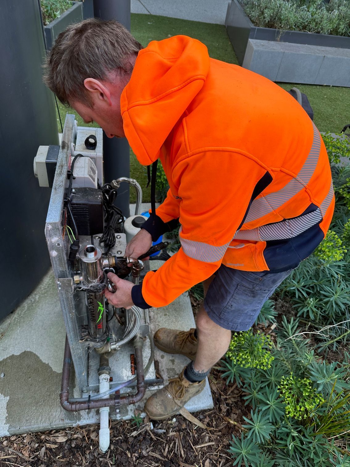 Worker in bright orange hoodie repairs machinery outdoors beside water — PUMPtek in Grovedale, VIC