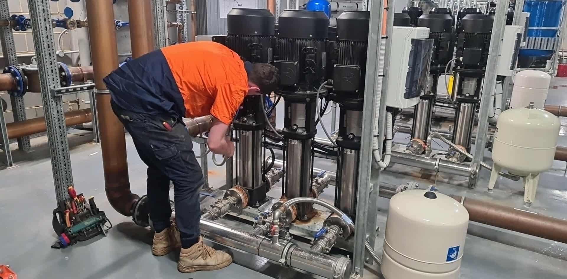 Worker in orange shirt bending over industrial piping and tanks in a mechanical room  — PUMPtek in Bendigo, VIC