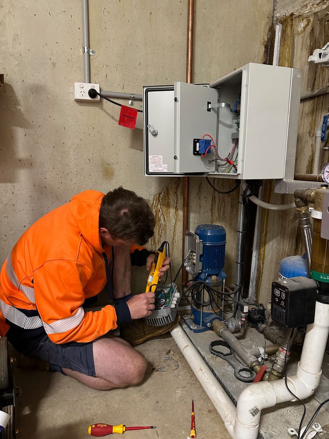 Technician in orange jacket kneels by a boiler, inspecting exposed pipes and electrical controls — PUMPtek in Bendigo, VIC