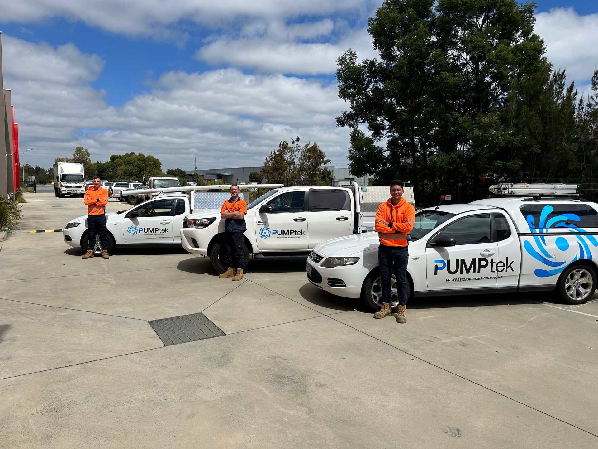 Three orange-clad workers stand beside white PUMPtex service vans in an industrial lot — PUMPtek in Grovedale, VIC