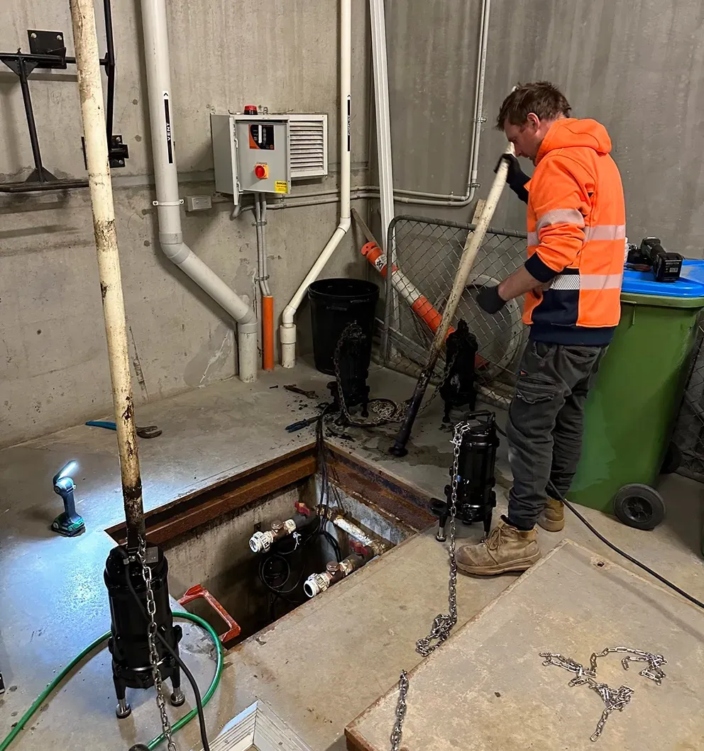 Worker in orange jacket inspecting a sewer pit with pipes and equipment in an industrial room — PUMPtek in Grovedale, VIC