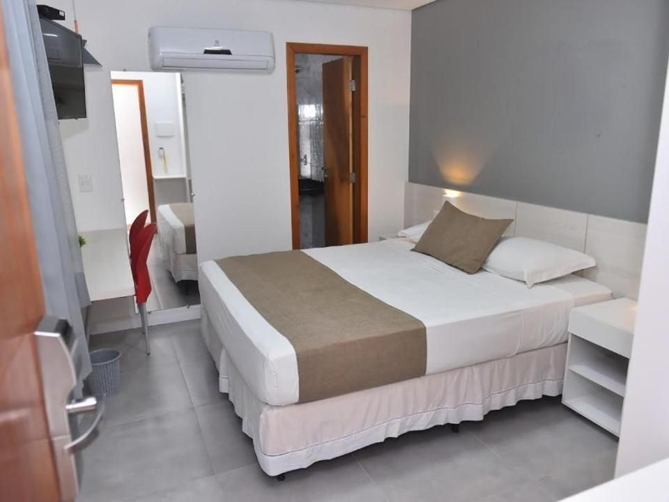 Um quarto de hotel com uma cama com edredom branco e peseira marrom, uma parede com detalhes em cinza e uma porta com moldura de madeira.